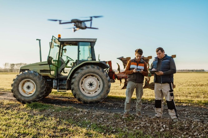 Imaginea prezintă două persoane aflate într-un câmp, lângă un tractor mare de culoare verde. Deasupra lor zboară o dronă, iar cei doi par să o controleze sau să utilizeze dispozitive electronice. Fundalul este format dintr-un câmp deschis, sub un cer senin.