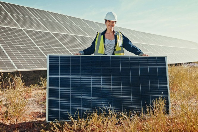 Imaginea prezintă o persoană purtând o cască de protecție albă și o vestă reflectorizantă, aflată în fața unui panou solar de mari dimensiuni. Persoana ține panoul cu ambele mâini. În fundal se pot observa mai multe rânduri de panouri solare montate pe sol, într-un câmp deschis cu iarbă și vegetație. Cerul este senin și albastru, sugerând o zi însorită.