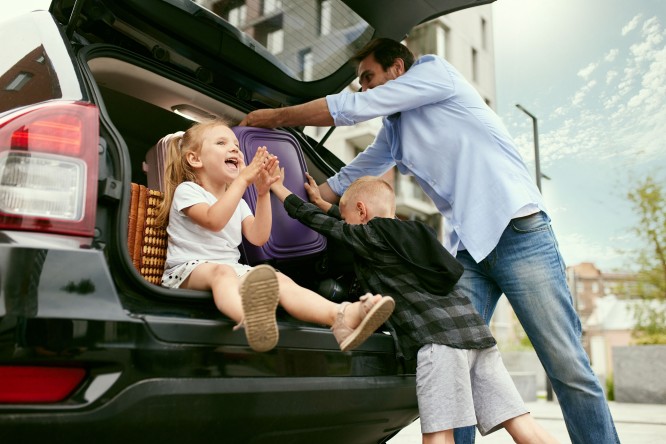 Imaginea prezintă un bărbat și doi copii în spatele unui portbagaj deschis de mașină. Bărbatul așază o valiză mov în portbagaj, în timp ce unul dintre copii, aflat în interiorul portbagajului, bate palma cu celălalt copil care stă în exterior. Fundalul sugerează un cadru urban, cu clădiri și copaci vizibili.