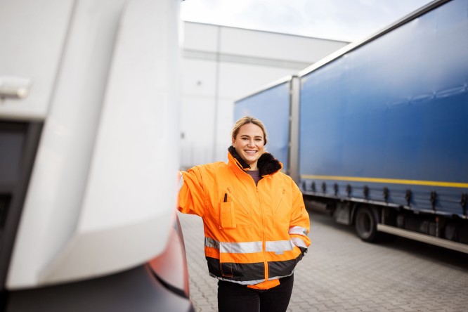 Imaginea prezintă o persoană îmbrăcată într-o jachetă portocalie cu elemente reflectorizante, aflată lângă un camion alb. În fundal se observă o remorcă albastră, iar scena are loc într-o zonă industrială sau într-un spațiu de depozitare, cu suprafață asfaltată și o clădire mare în spate.