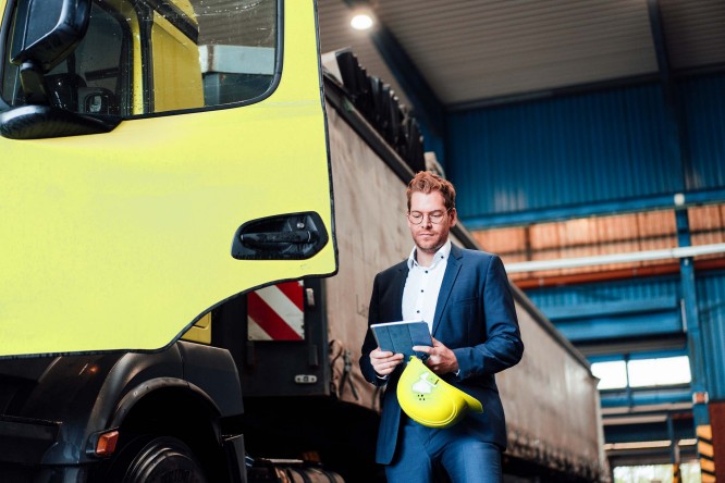 Imaginea prezintă o persoană îmbrăcată în costum, care ține în mână o tabletă și o cască de protecție galbenă, aflată lângă un camion mare de culoare galbenă, într-un depozit industrial. Spațiul are pereți albaștri și structuri metalice, sugerând un mediu de lucru industrial sau logistic.