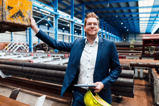 Imaginea prezintă o persoană îmbrăcată într-un costum albastru, aflată într-un spațiu industrial — posibil un depozit sau o fabrică. Aceasta ține în mână o cască de protecție galbenă și o tabletă sau clipboard. În fundal se observă țevi metalice, grinzi și diverse echipamente industriale, majoritatea vopsite în albastru. Persoana pare să fie implicată într-un proces de inspecție sau supraveghere a activităților din unitate.