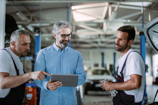 Imaginea prezintă trei persoane într-un atelier auto sau garaj. Două dintre ele poartă salopete de lucru, iar una ține în mână o cârpă. Persoana din mijloc poartă o cămașă albastră deschisă și ține o tabletă, în timp ce persoana din stânga pare să indice ceva pe ecranul acesteia. În fundal se observă diverse unelte și echipamente specifice unui service auto, inclusiv un elevator pentru mașini și un vehicul.